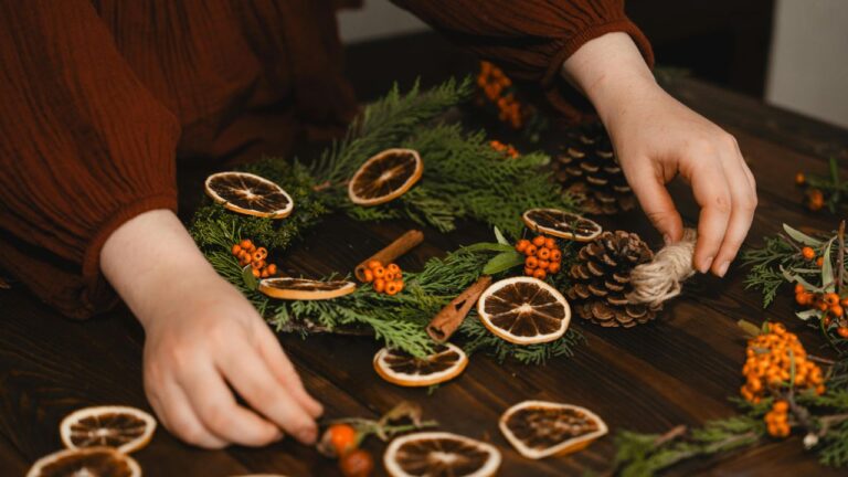 Hands creating a winter wreath with dried oranges, pine, cedar, berries, and pinecones for a December witchcraft ritual.